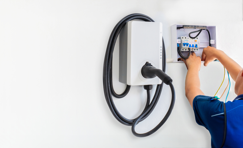 Person installing or wiring an electric vehicle charging station on a white wall, with cables connected to a junction box.