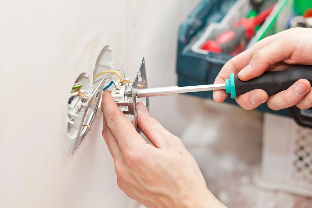 A person uses a screwdriver to install or repair an electrical outlet, connecting wires inside a wall. A toolbox with tools is visible in the background.