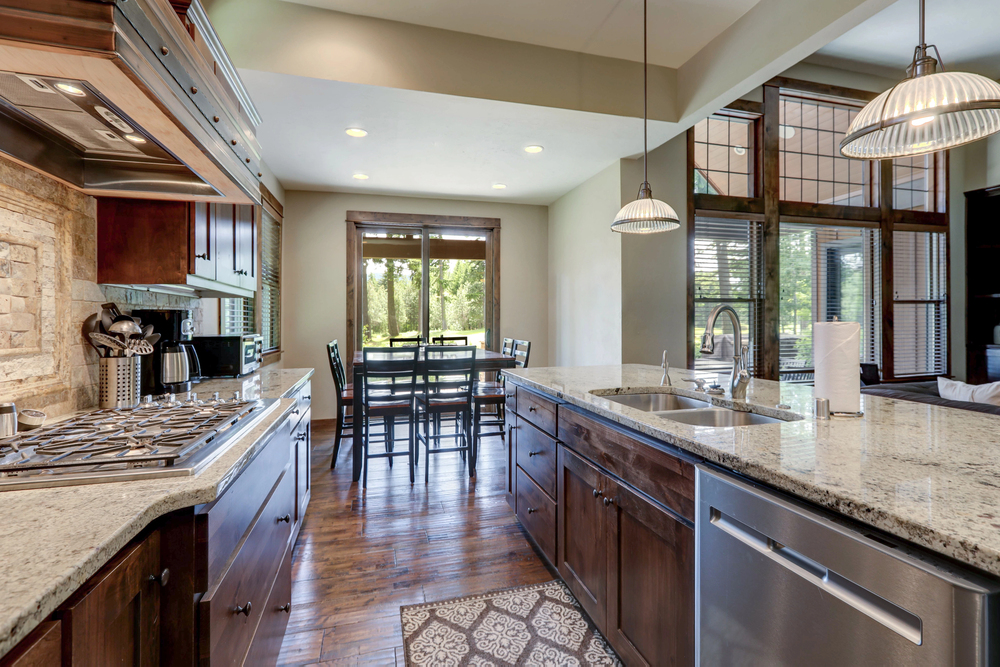 Modern kitchen with granite countertops, stainless steel appliances, wooden cabinets, and a dining area with large windows showing greenery outside.