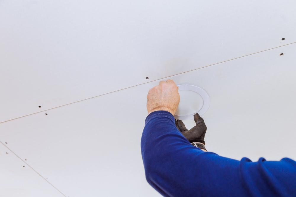 Person wearing a blue long-sleeve shirt and black glove installing or adjusting a round recessed ceiling light fixture on a white ceiling.